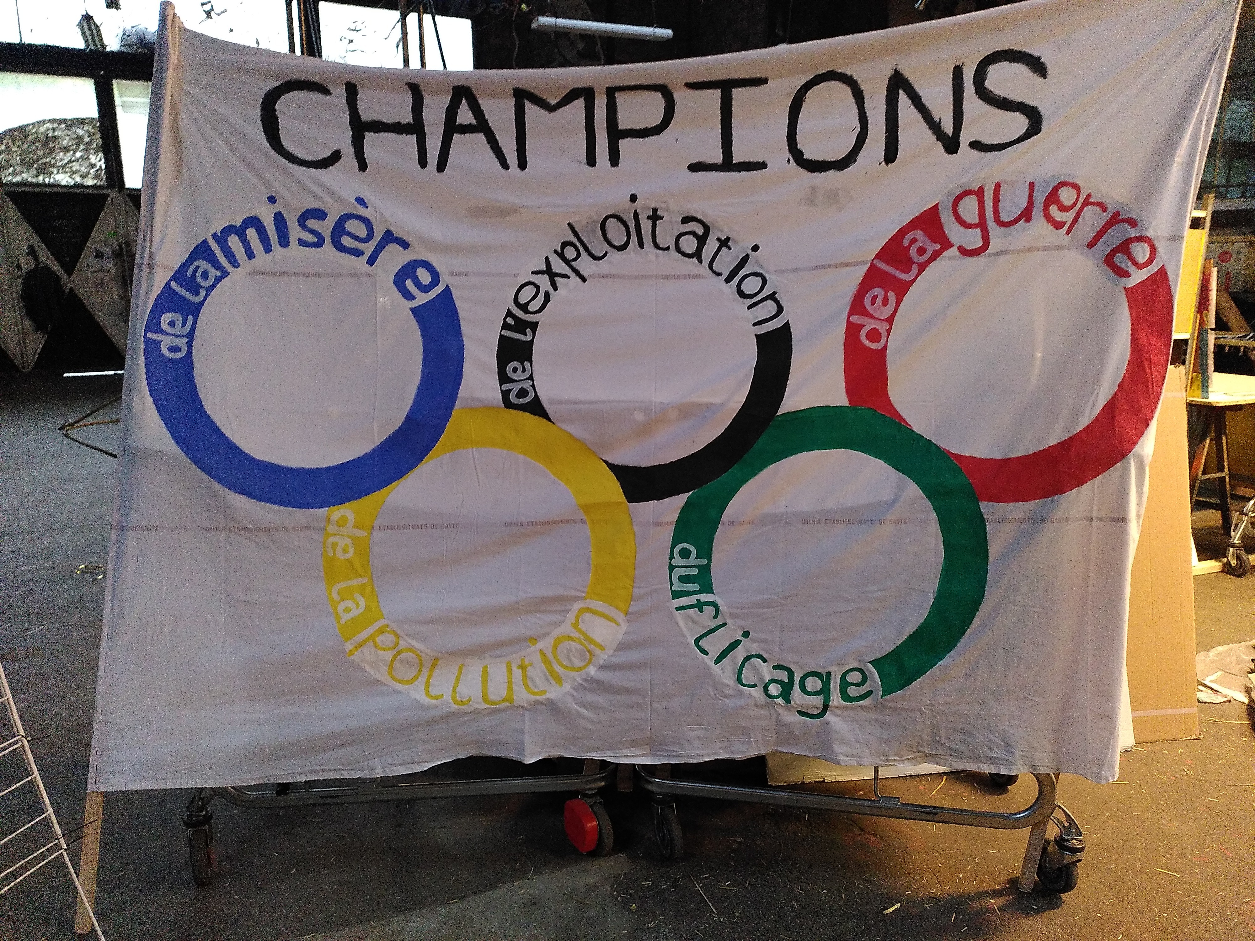Banderole anti-JOP, un texte Champions surplombe les anneaux olympiques portant les mentions: de la misère, de de l'exploitation, de la guerre, de la pollution, du flicage 